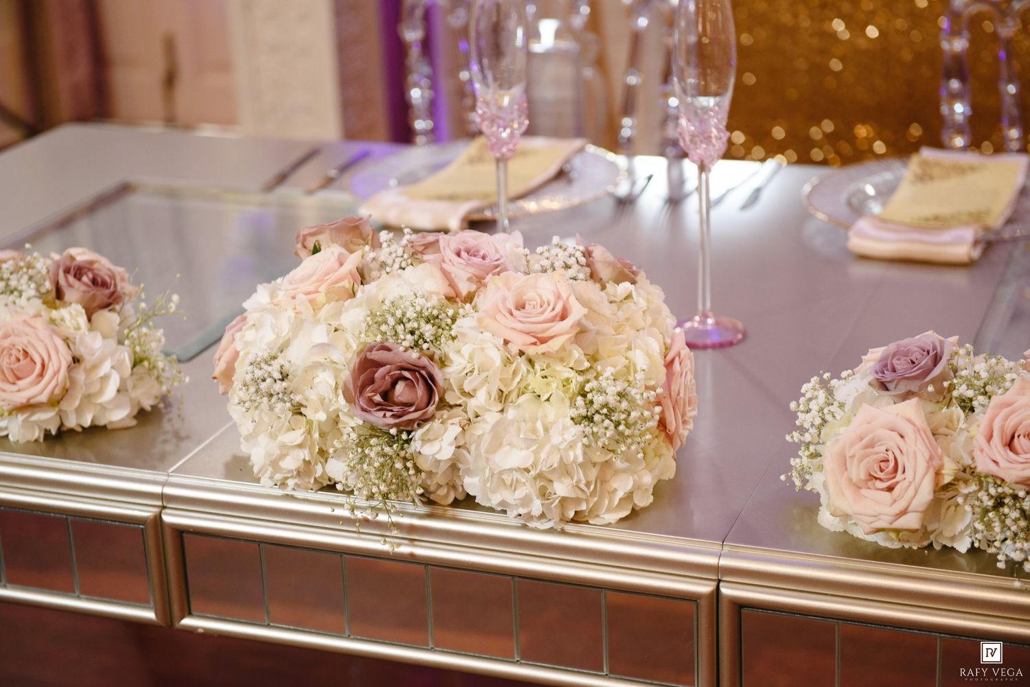 Elegant wedding table centerpiece with blush pink roses, white hydrangeas, and baby's breath on a mirrored surface.
