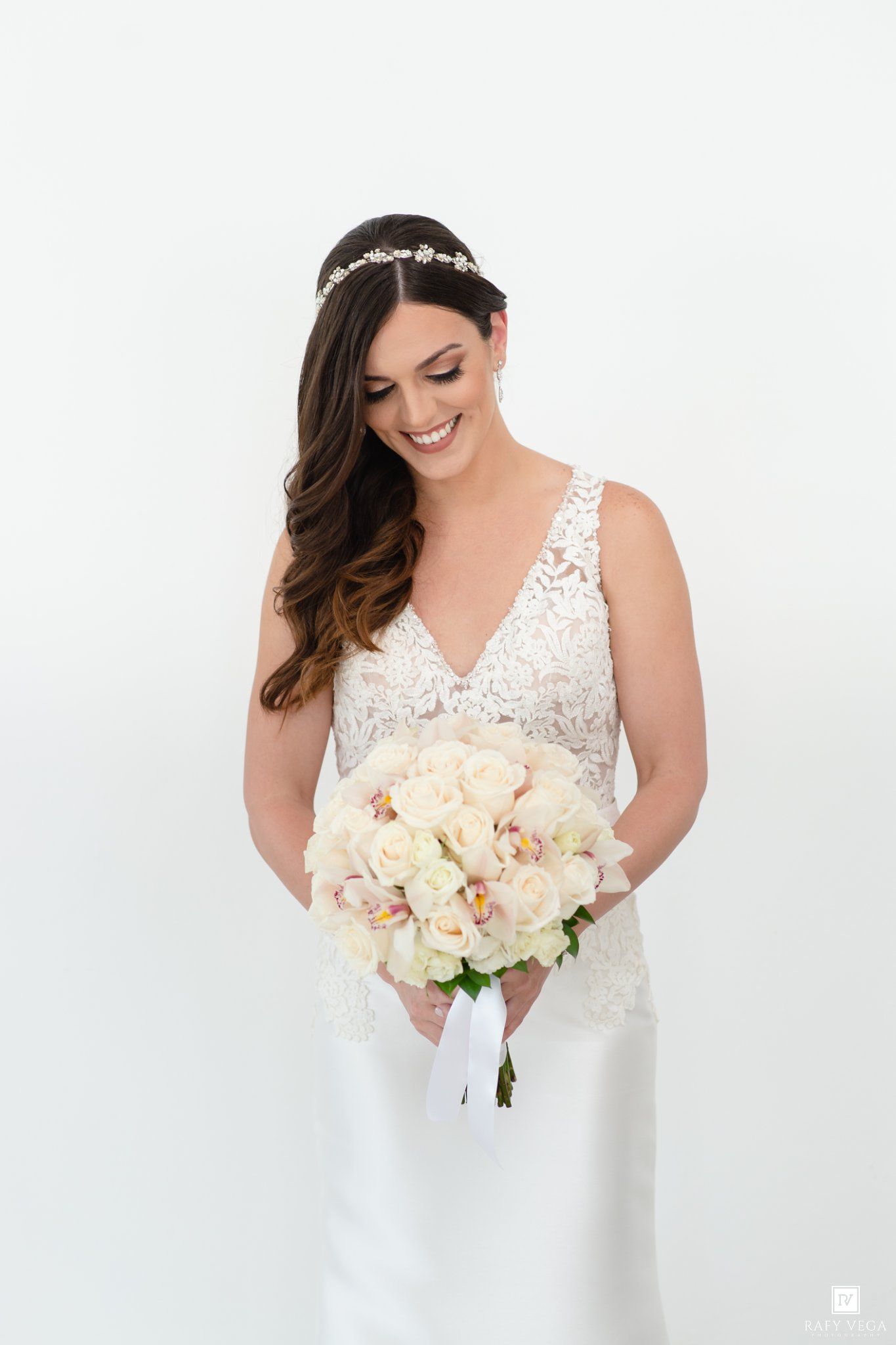Smiling bride in lace V-neck gown holding white and blush rose bouquet with floral headpiece against white background.