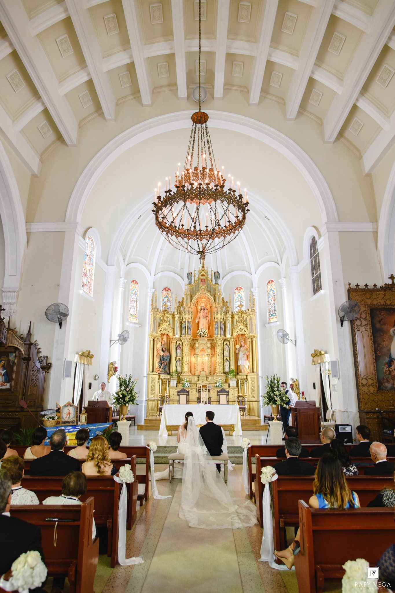 Elegant wedding ceremony in a white Catholic church with ornate golden altar, chandelier, and bride and groom at the altar.