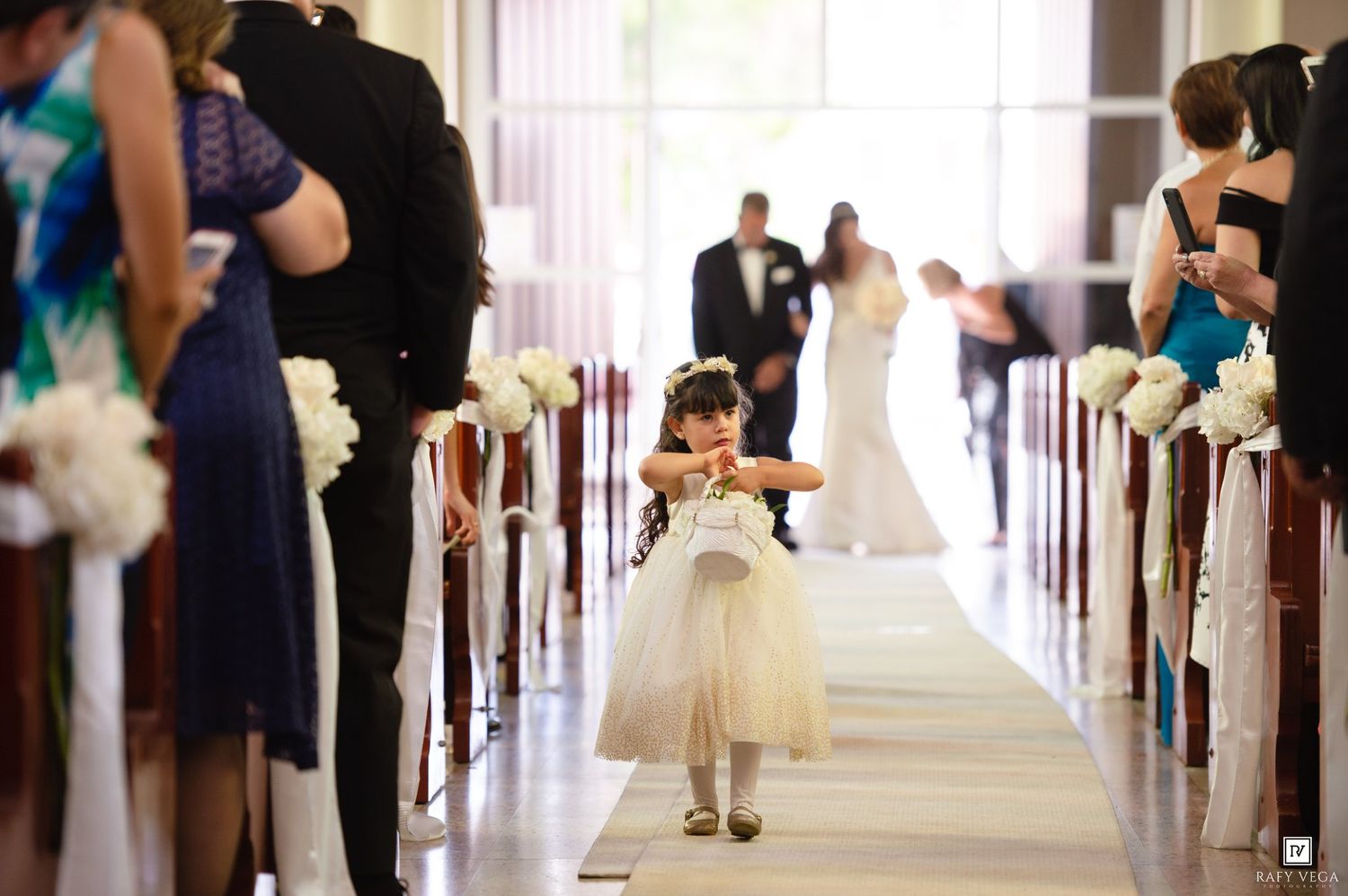 Flower girl in white dress walks down church aisle during wedding ceremony with bride and groom in background.