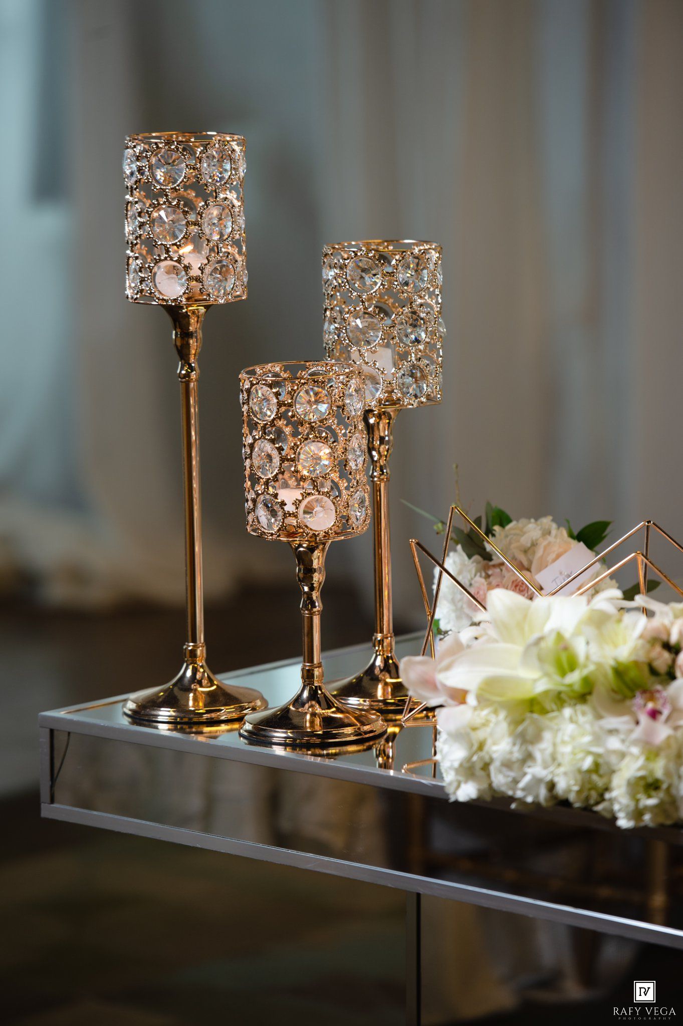 Gold crystal candleholders of varying heights on a mirrored tray beside a white floral arrangement at an elegant event.
