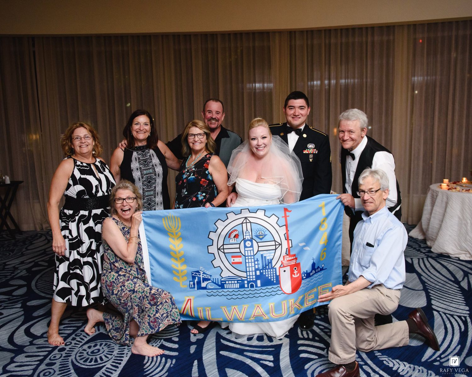 Wedding group photo with guests holding a Milwaukee city flag at an indoor reception venue.