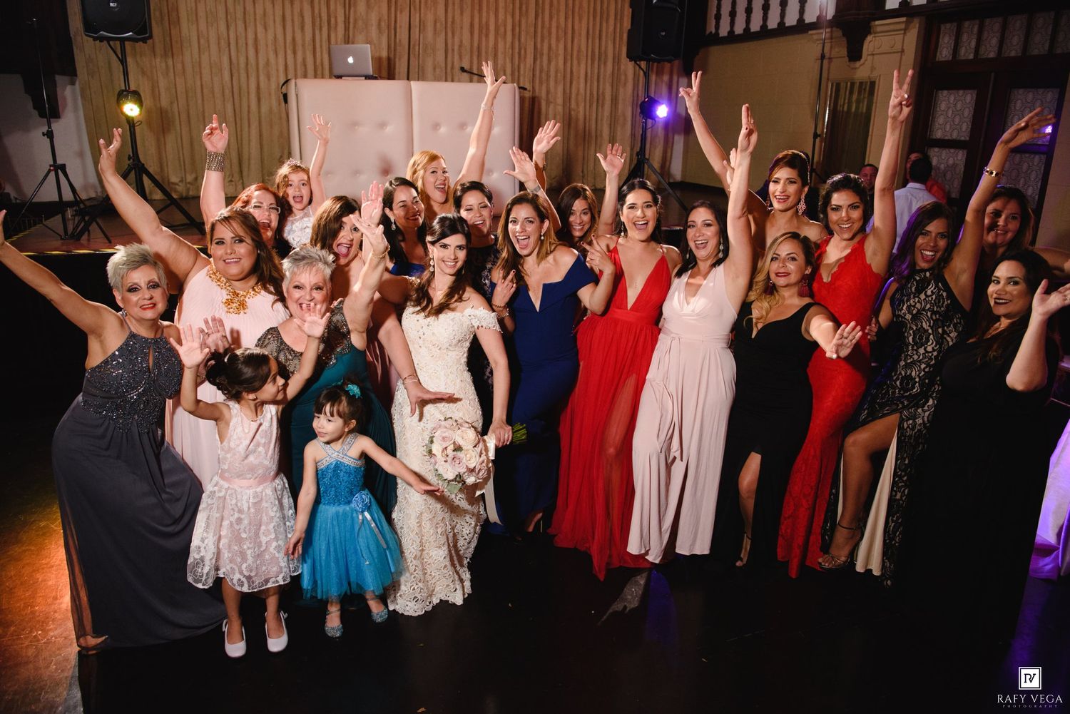 Bride and female guests celebrate together on the dance floor at a wedding reception, arms raised in joy.