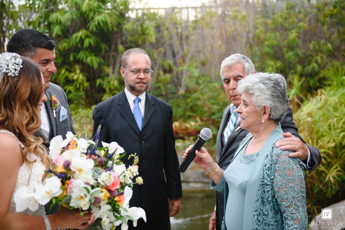 Elderly woman speaks at outdoor wedding ceremony with bouquet, officiant, and guests in garden setting.