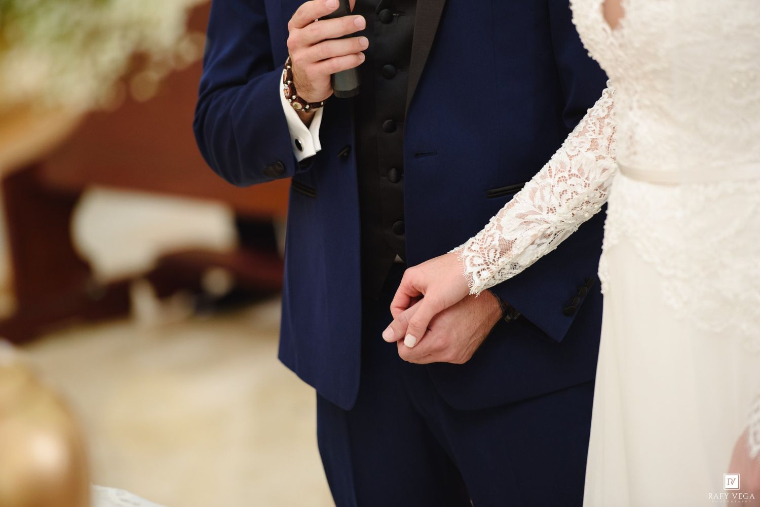 Bride and groom holding hands at wedding ceremony, groom in navy blue suit, bride in lace white gown.