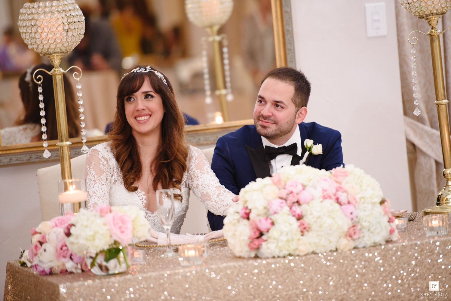 Bride and groom smiling at sweetheart table with pink floral arrangements and gold candelabras at elegant wedding reception.