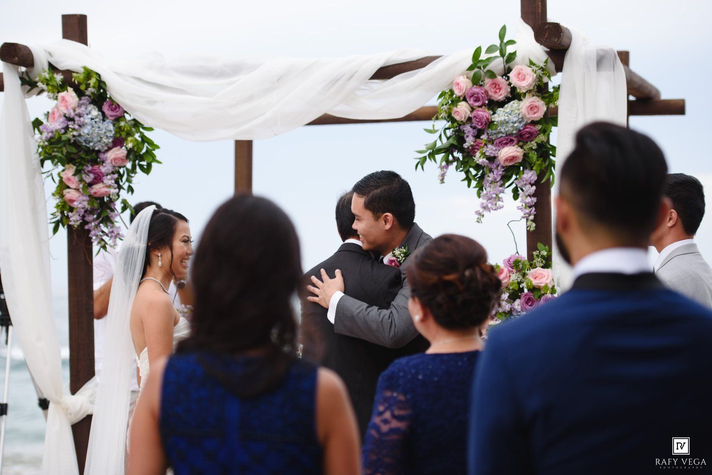 Bride and groom share a tender moment at an outdoor wedding ceremony under a floral arch with pink and white blooms.