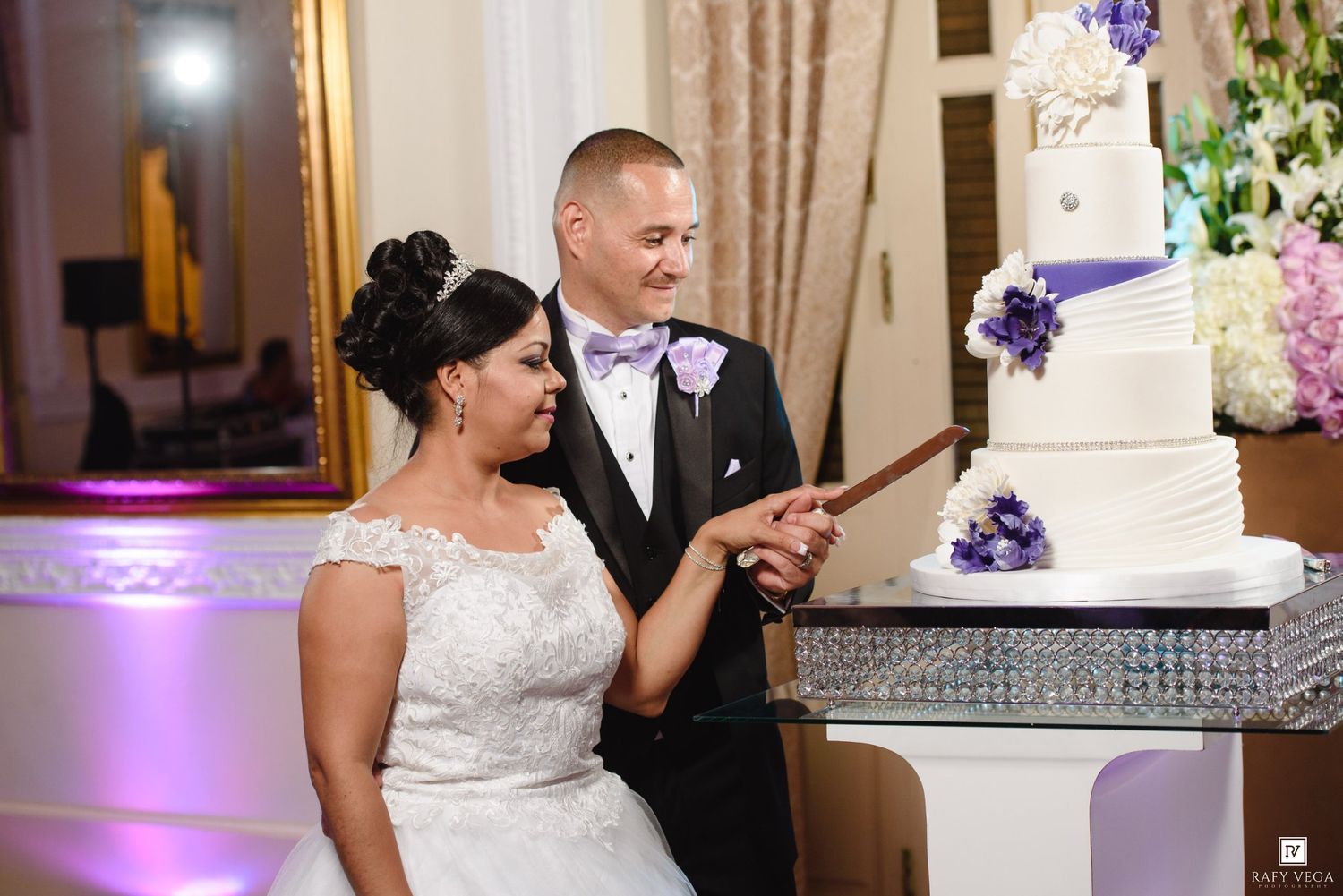 Bride and groom cutting their elegant white and purple tiered wedding cake at their reception venue.