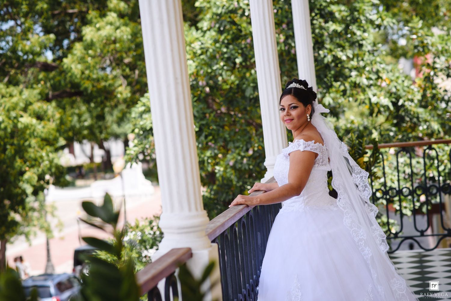 Bride in white gown and veil poses gracefully on an elegant balcony with white columns and lush green trees.