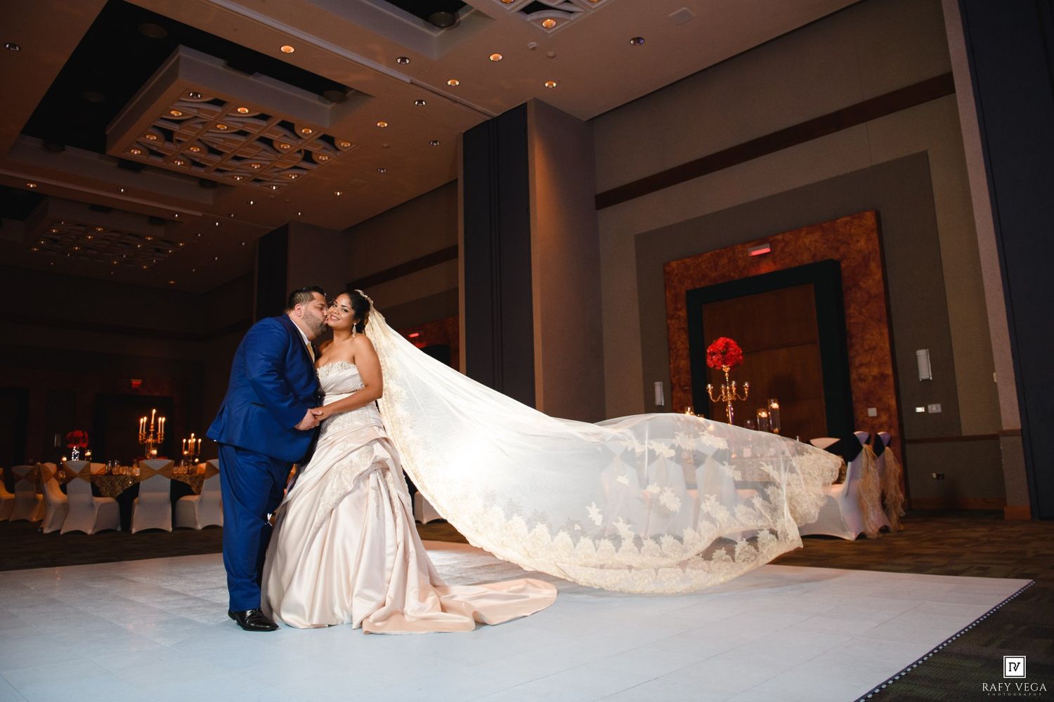 Bride and groom share a romantic kiss on white dance floor, her long flowing veil trailing elegantly behind her.