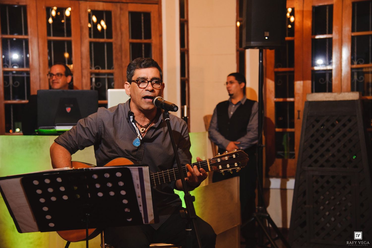 Musician playing acoustic guitar and singing into microphone at indoor evening event with band members in background.