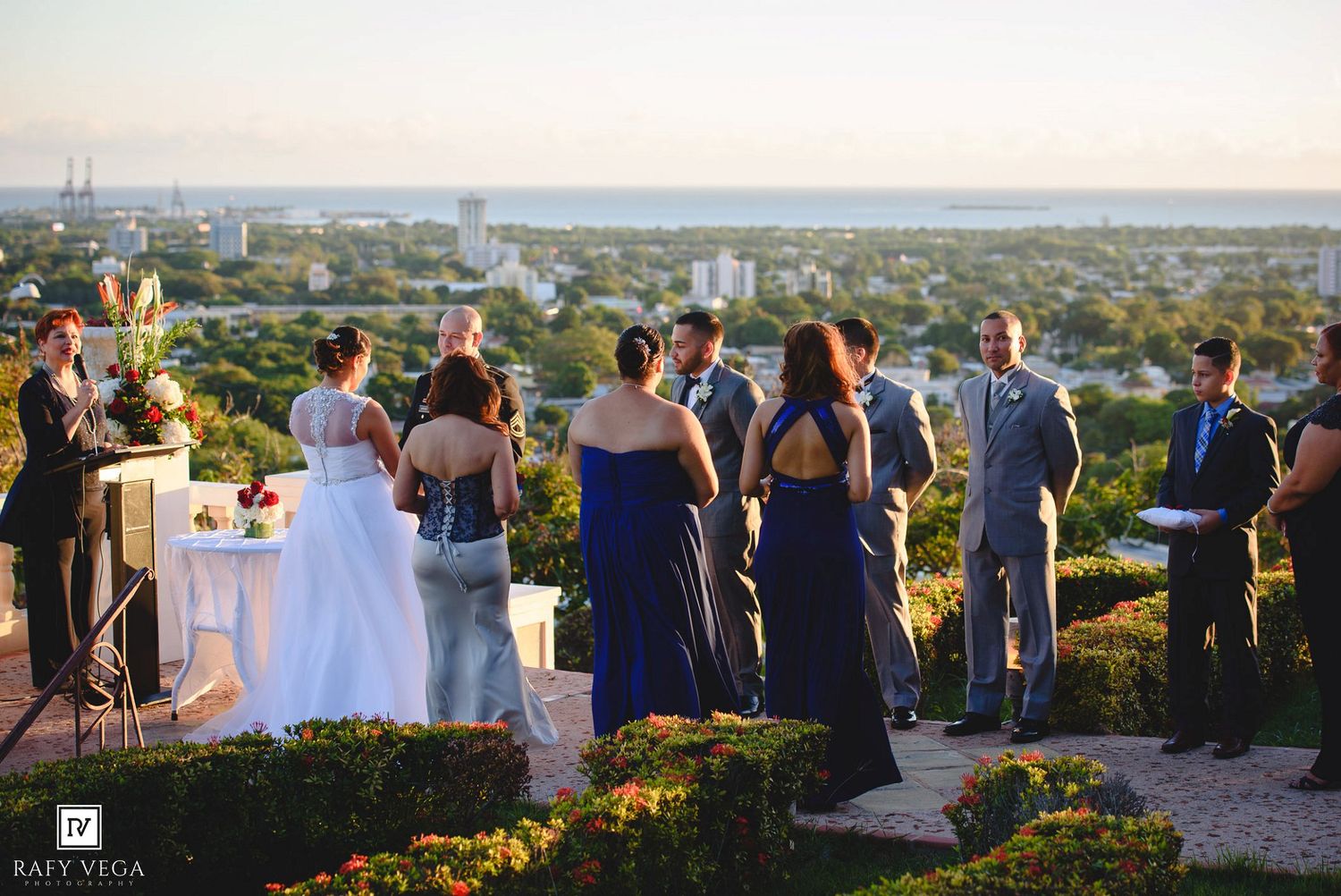 Outdoor wedding ceremony on a hilltop terrace with bridal party in navy blue, overlooking a scenic city and waterfront view.
