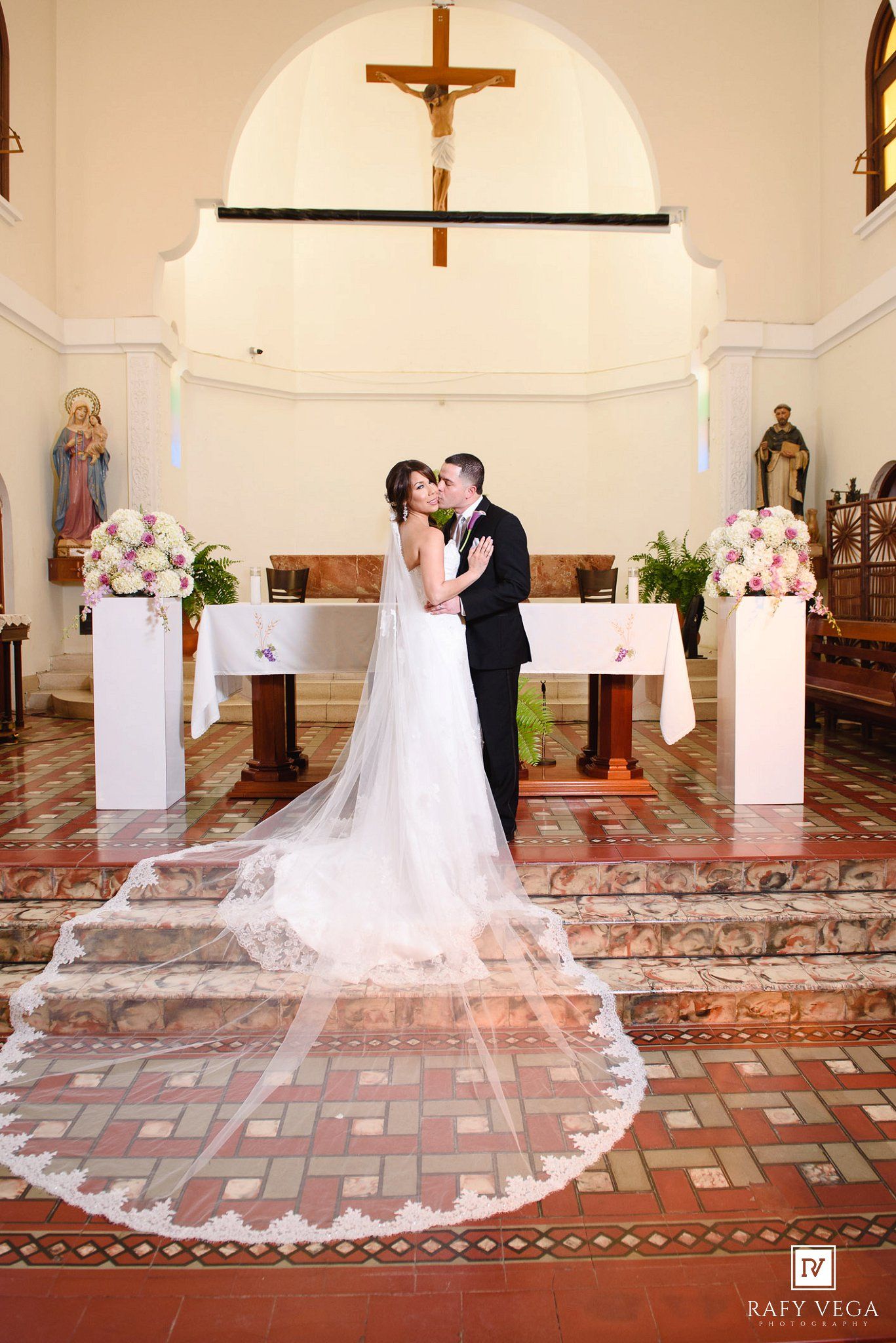 Bride and groom share first kiss at church altar with long lace veil trailing.