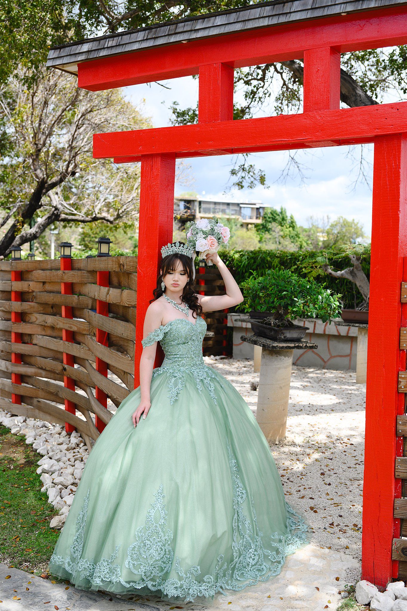 Young woman in sage green quinceañera ball gown with tiara poses under red torii gate in garden.