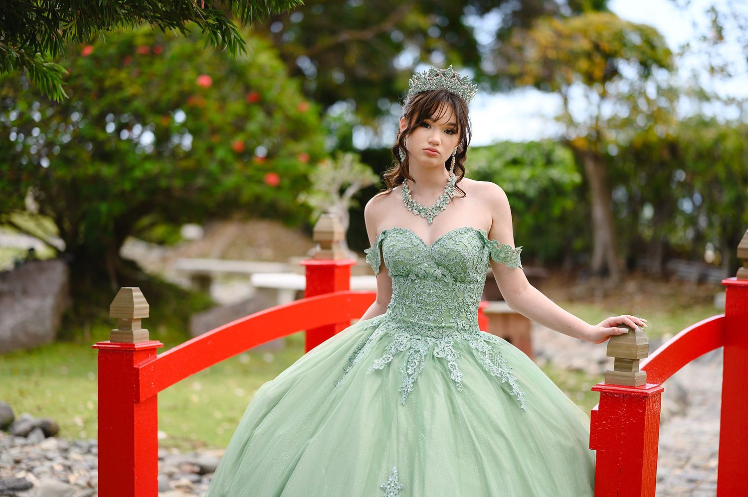 Young woman in mint green ball gown with lace details and tiara poses gracefully by red garden bridge outdoors.