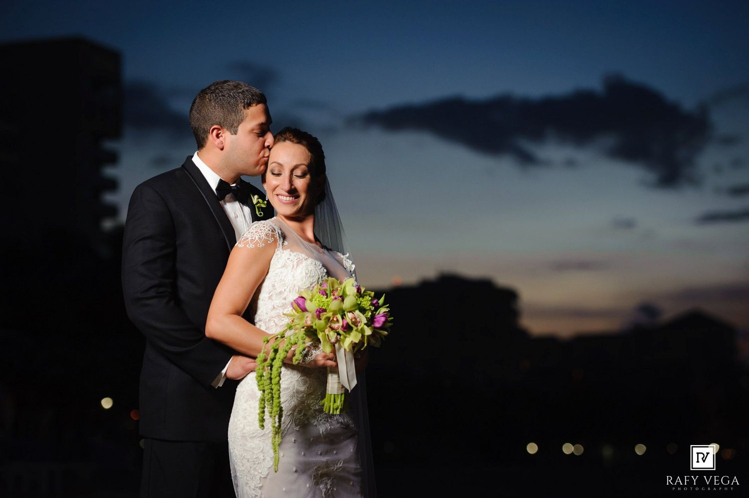 Bride and groom share a romantic moment at dusk, bride holding green floral bouquet, groom in black tuxedo kissing her cheek.