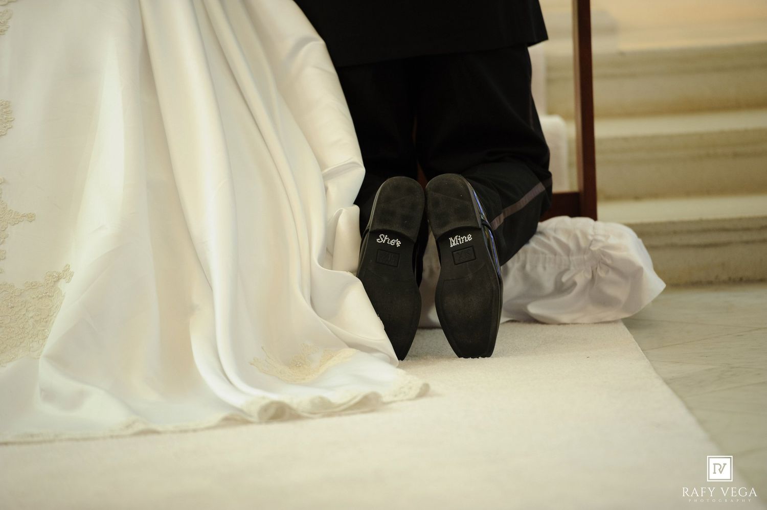 Wedding couple kneeling showing groom shoe soles with \u201cI do\u201d written on them, bride in white gown beside him.