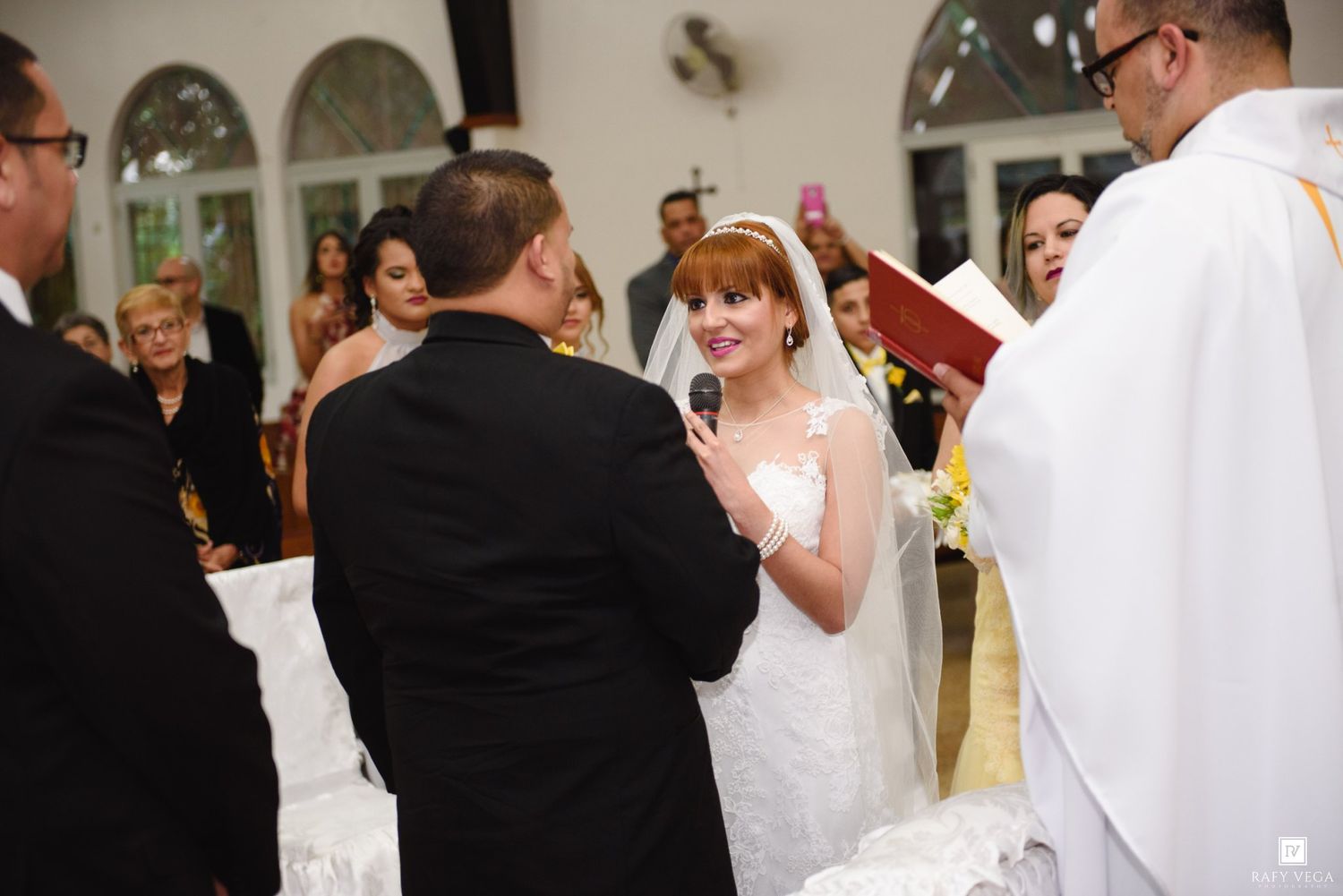Bride smiling and speaking into microphone during wedding ceremony at church altar with officiant and bridesmaids.