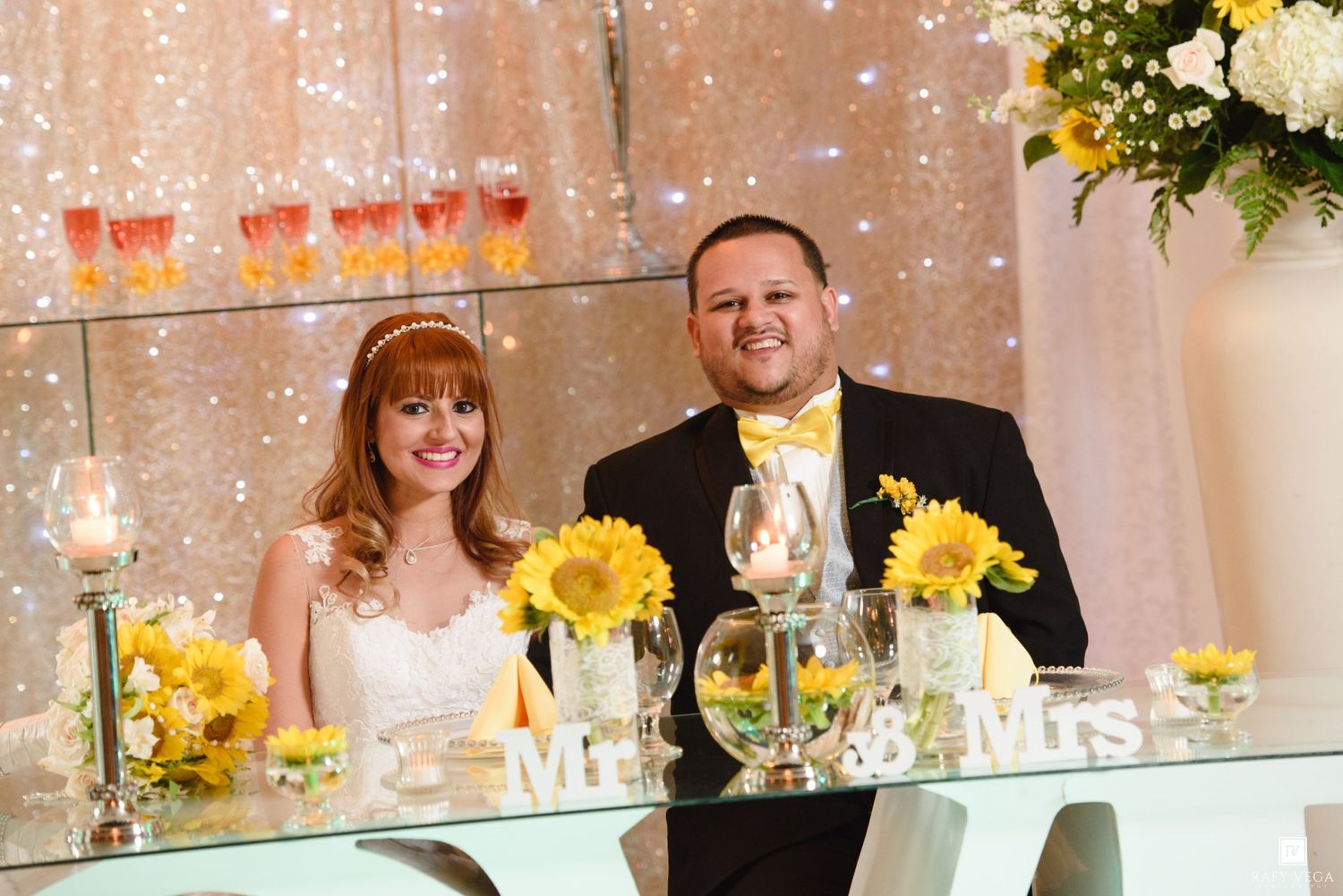 Bride and groom smiling at sweetheart table decorated with sunflowers and Mr. & Mrs. signs at wedding reception.