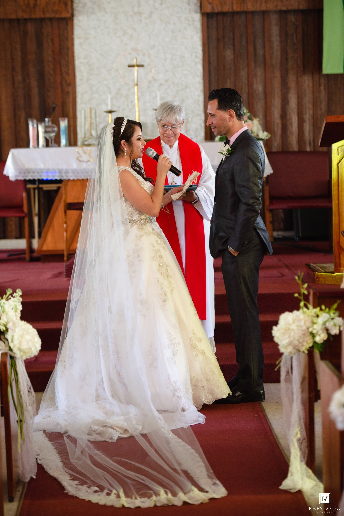 Bride and groom exchange vows at church altar with officiant in red vestments during elegant wedding ceremony.