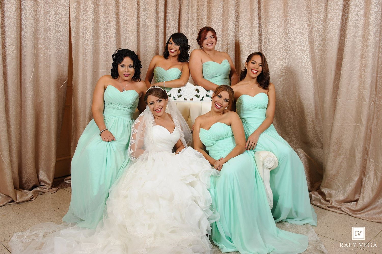 Bride in white gown seated with five bridesmaids in mint green dresses posing before a gold sequin backdrop.