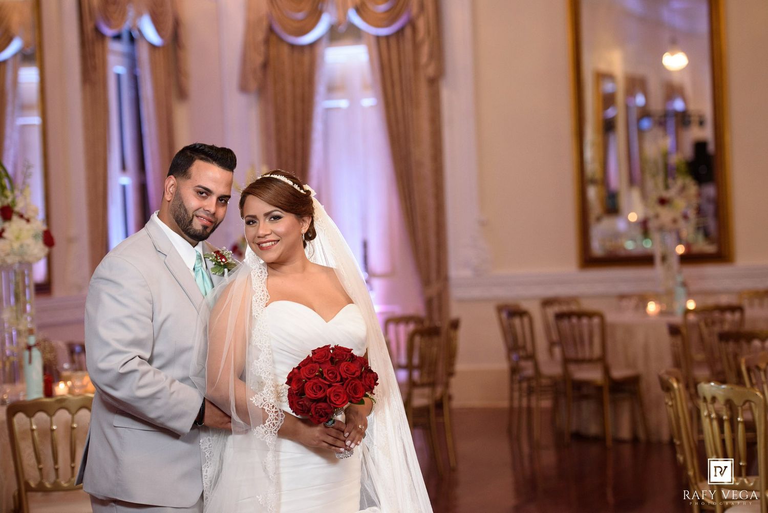 Bride and groom posing in elegant ballroom, bride holding red rose bouquet with veil, groom in gray suit with mint tie.
