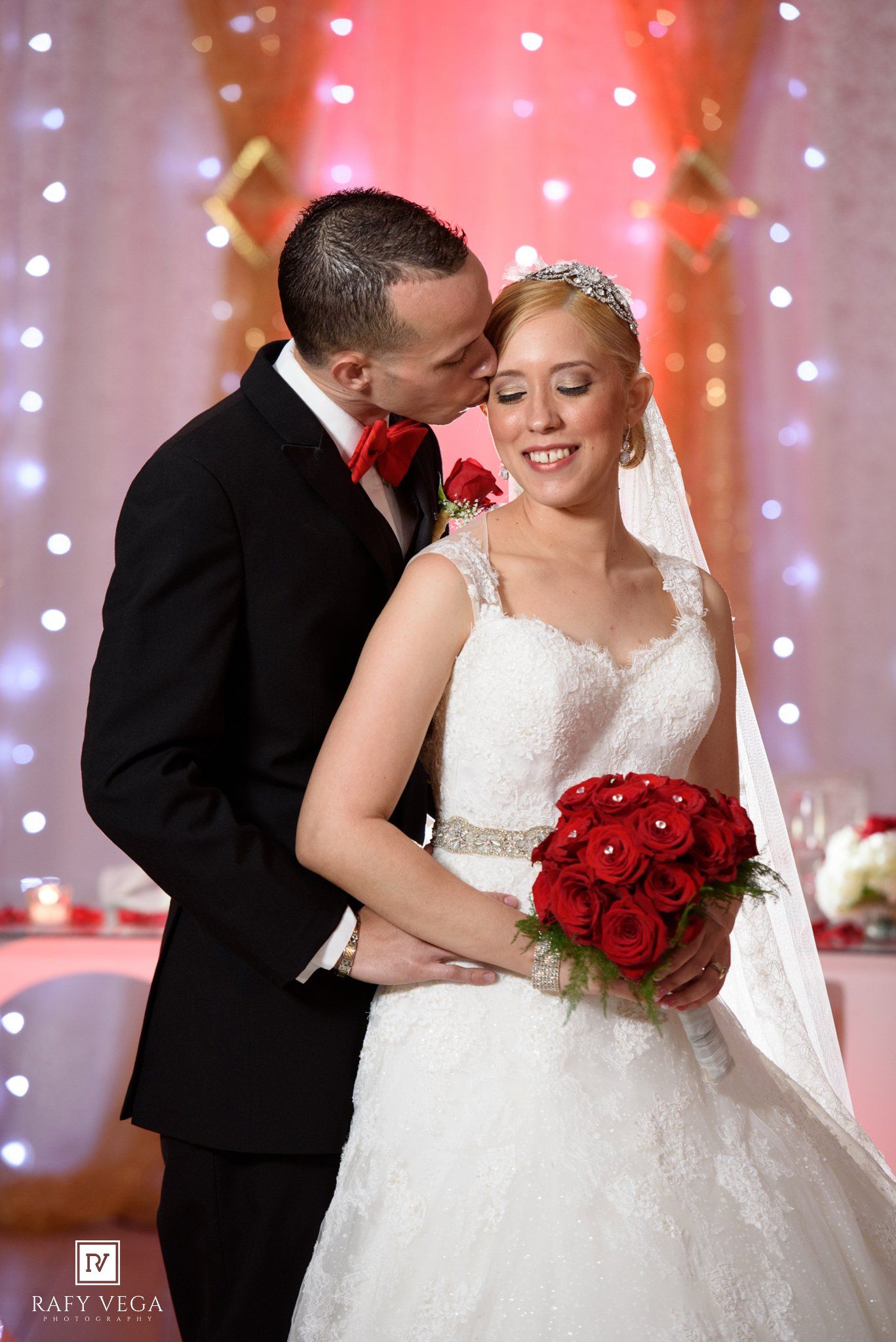 Bride in white lace gown holds red roses bouquet as groom in black tuxedo kisses her cheek at wedding reception.