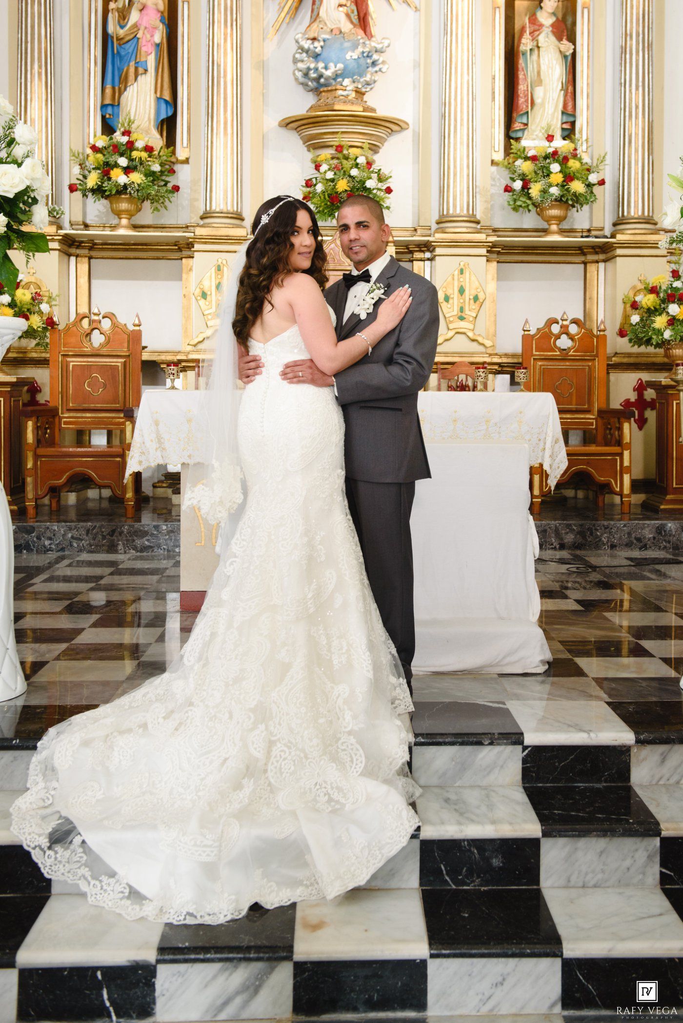 Bride in white lace gown and groom in gray suit pose together at ornate church altar with floral arrangements.