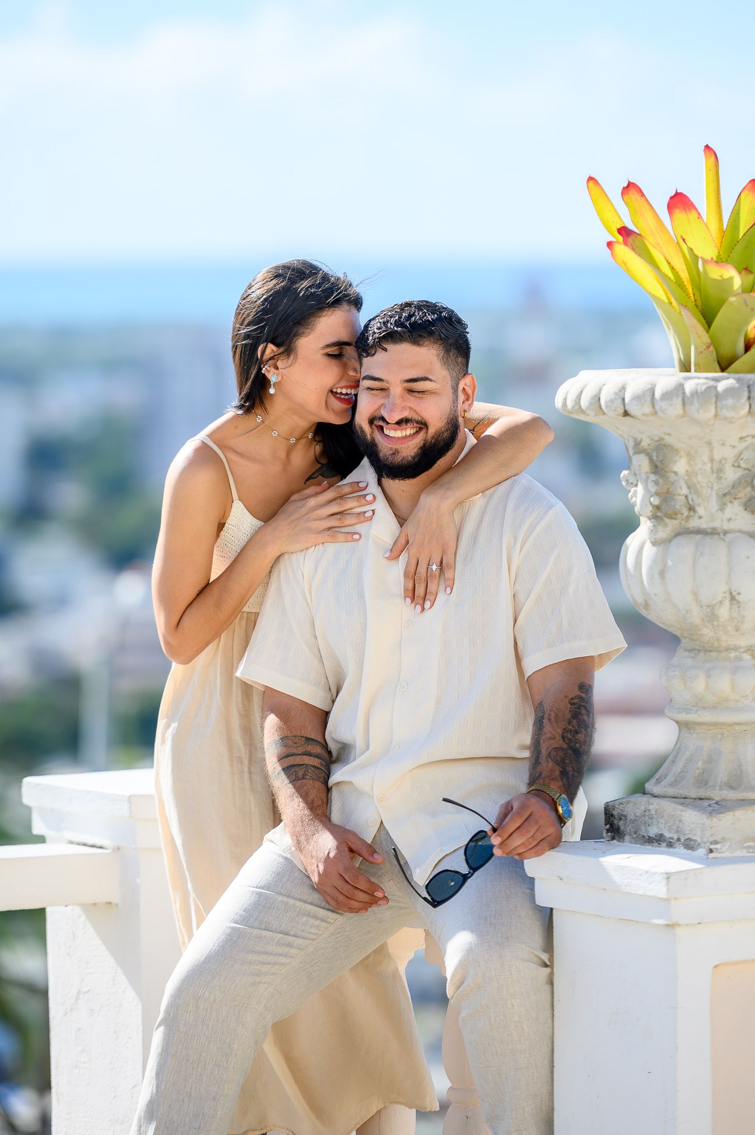 Smiling couple in cream outfits sitting together on white balcony with city view behind them.