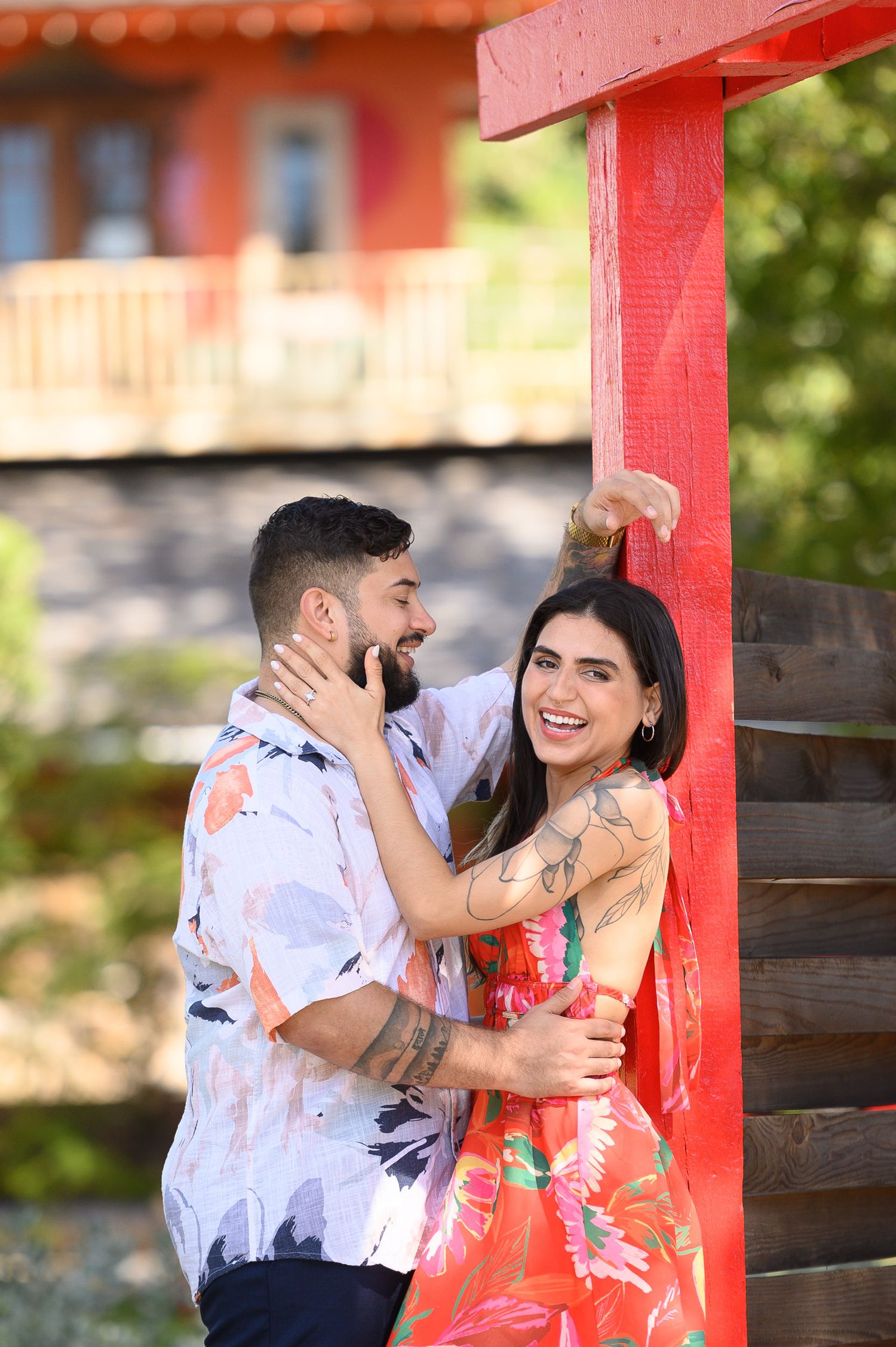 A smiling couple embraces by a red wooden post outdoors, woman in floral dress with tattoos, man in patterned shirt.