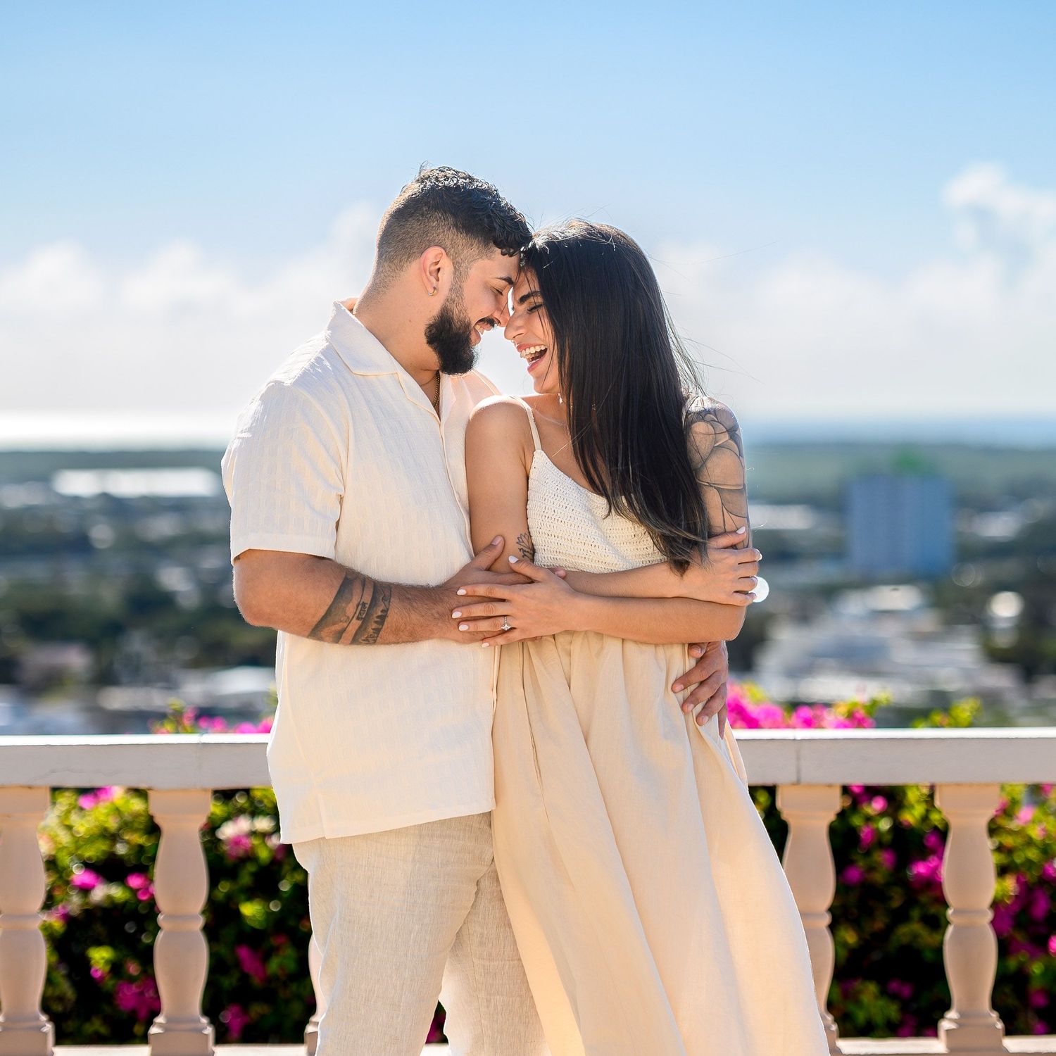 Couple embracing on a balcony with pink flowers and a scenic city view under a bright blue sky.