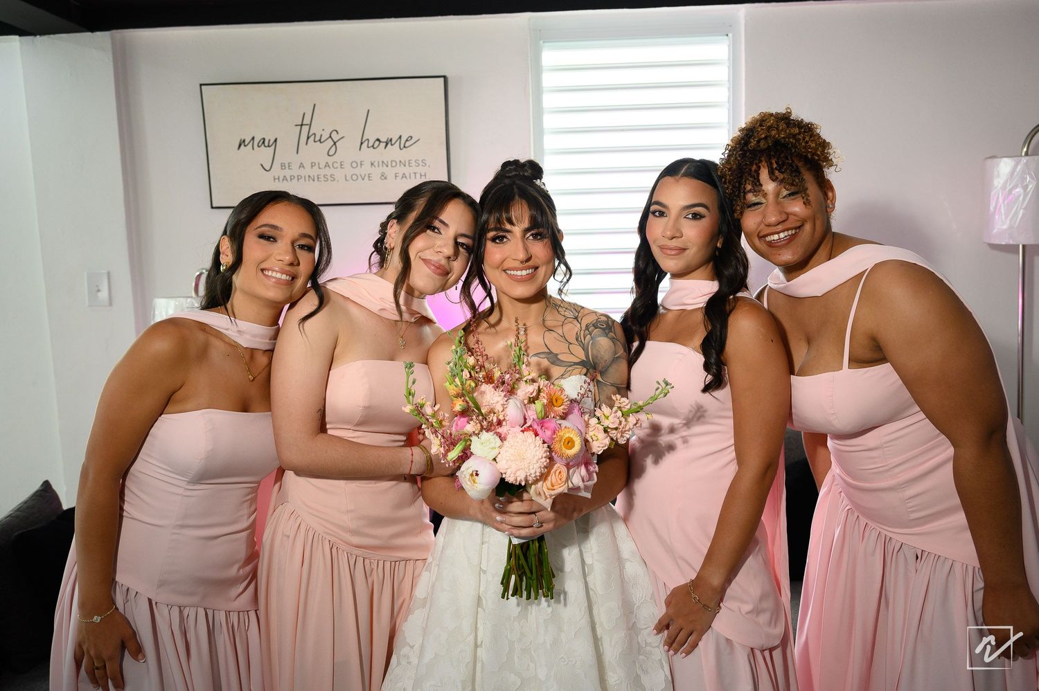Bride in a white gown holding a pink floral bouquet poses with four smiling bridesmaids in blush pink dresses in a bright room.