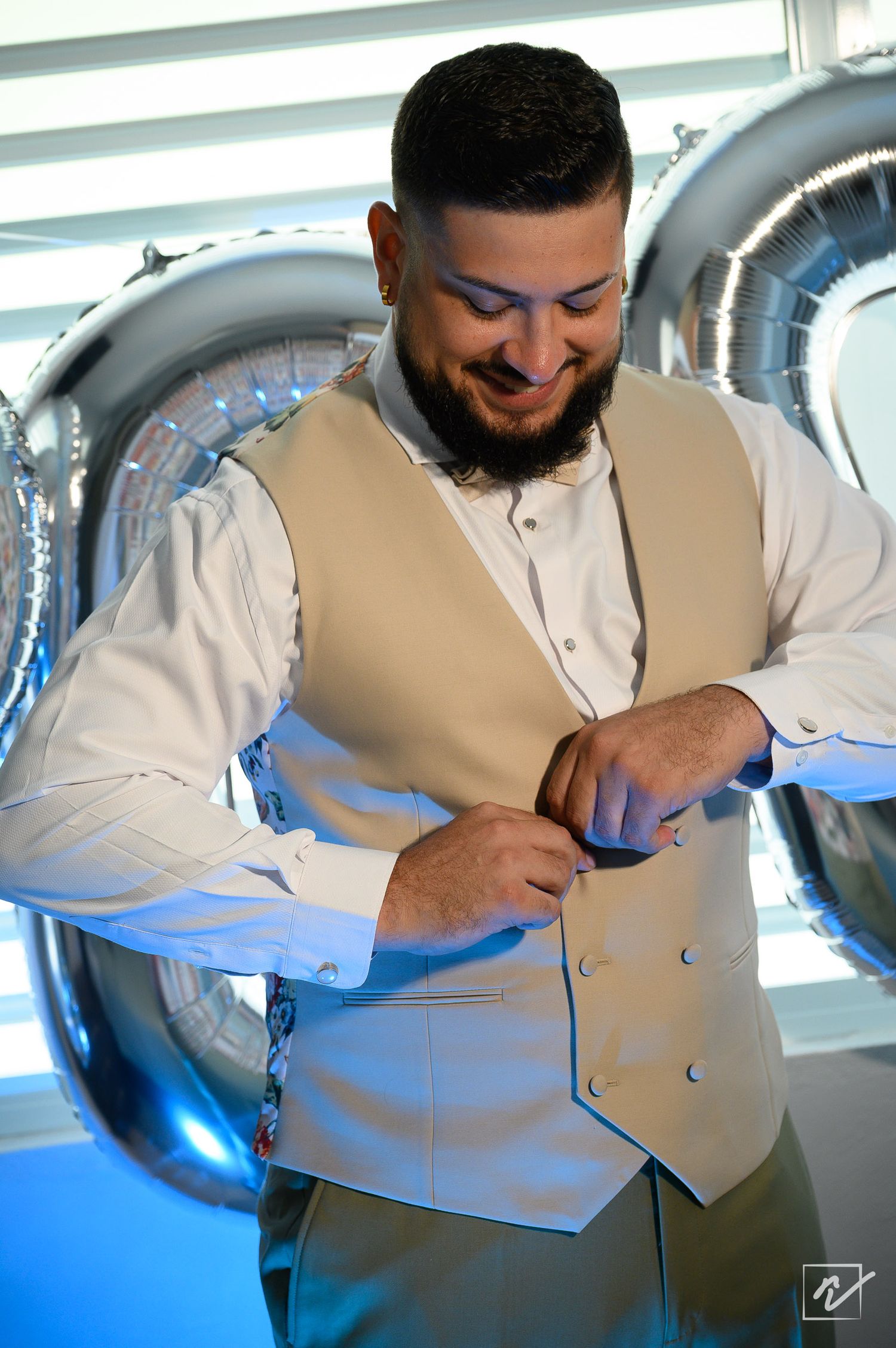 Smiling bearded groom in a tan vest and white dress shirt buttons his vest while getting ready beside large silver balloons.