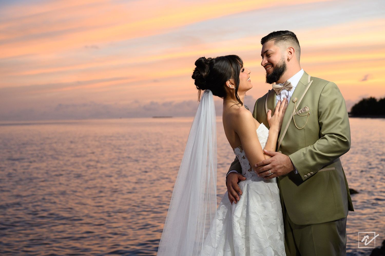 Bride in a white gown and veil embraces her bearded groom in an olive suit by the water at a colorful sunset wedding.