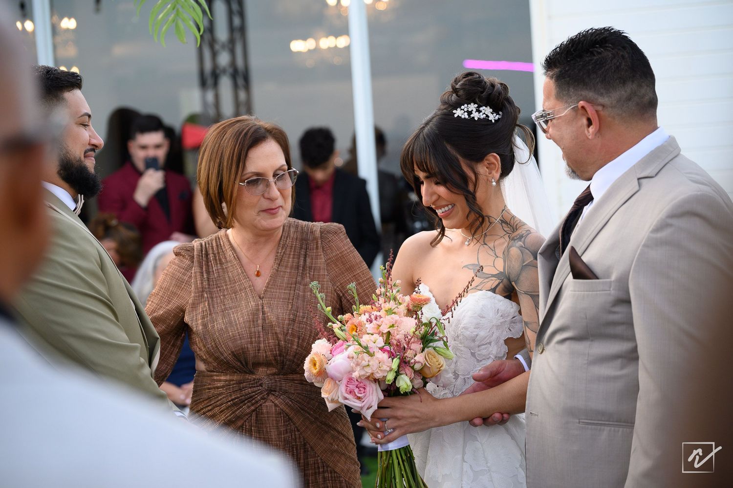 A smiling bride in a lace gown holding a pink floral bouquet embraces a woman in a bronze dress as a groom in a gray suit looks on.