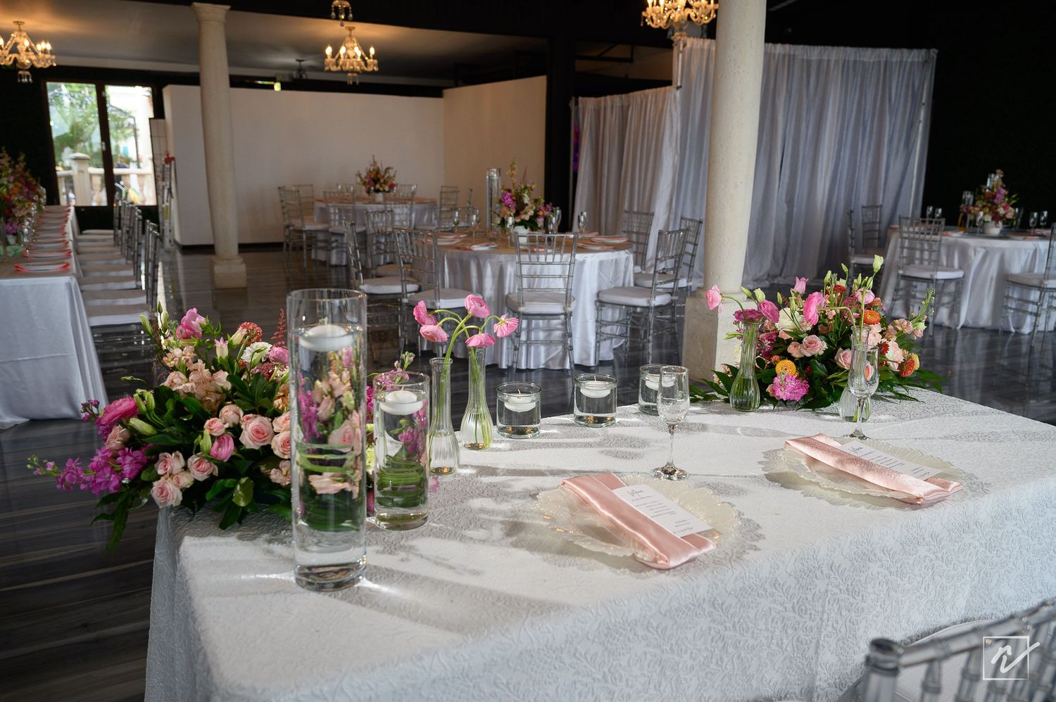 Elegant wedding reception table with pink floral centerpieces, glass vases, and pink napkins on white linen in a bright banquet hall.