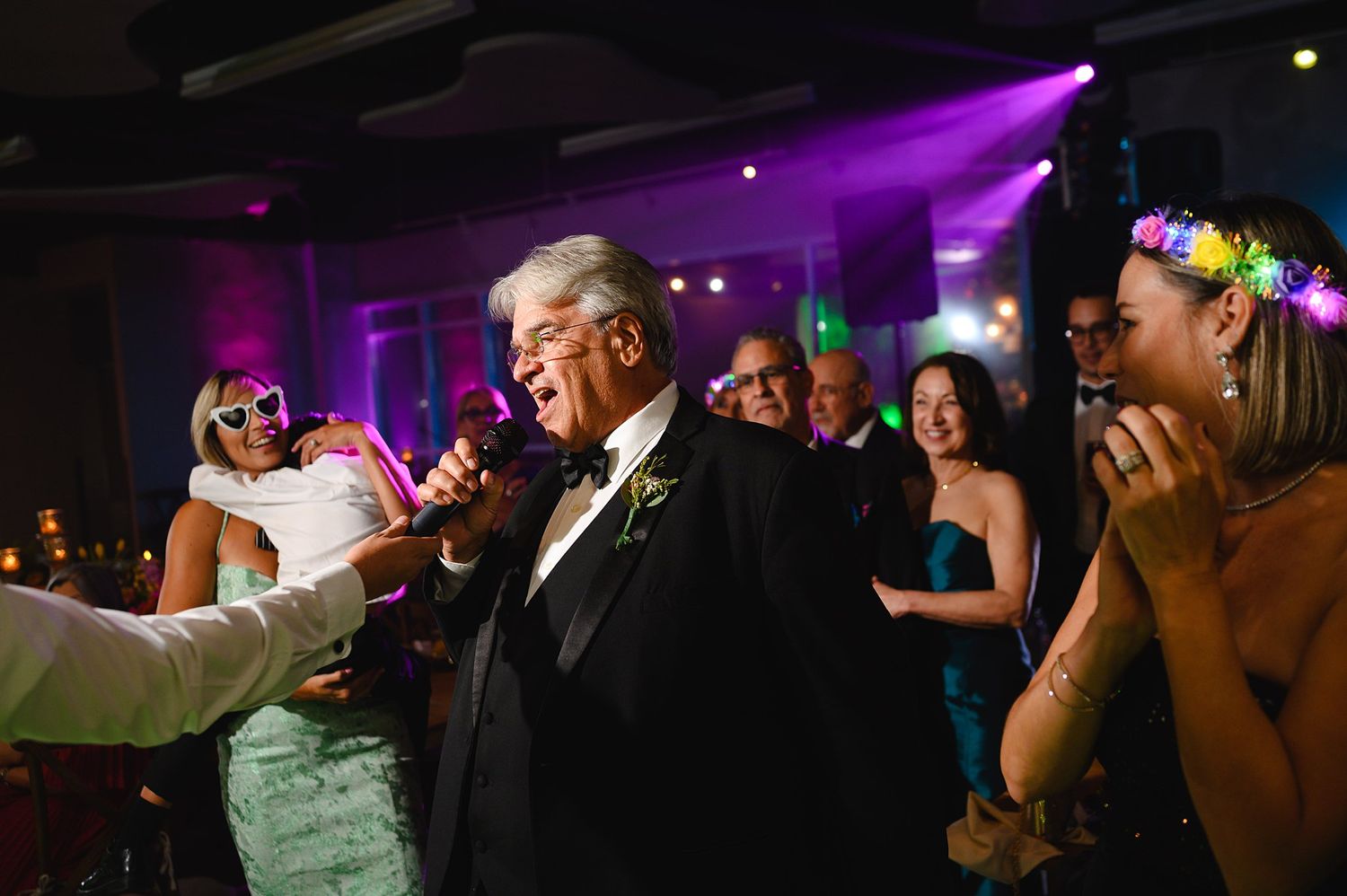 Elegant wedding reception with guests dancing, man in tuxedo singing with microphone under purple lights.