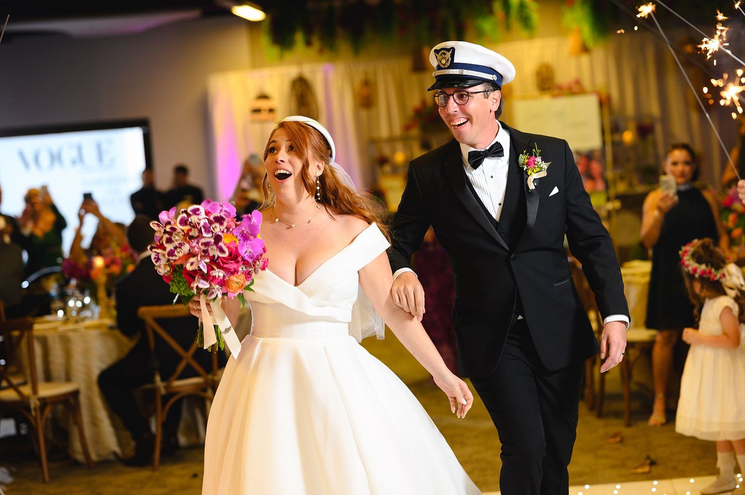 Bride in white gown and groom in tuxedo with captain hat walk joyfully at wedding reception with sparklers.