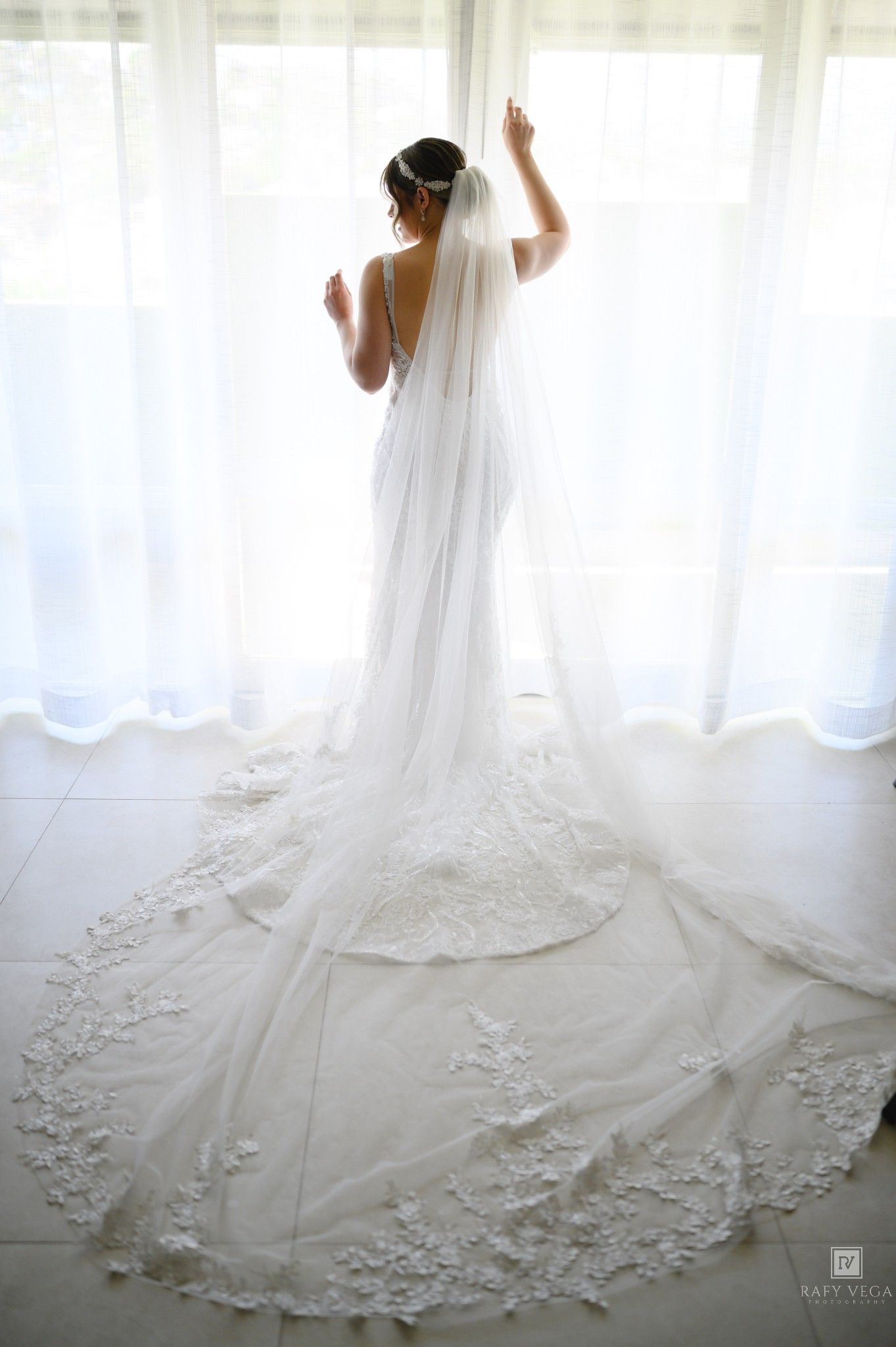 Bride in a stunning white lace wedding gown with a long cathedral veil stands by a bright window.