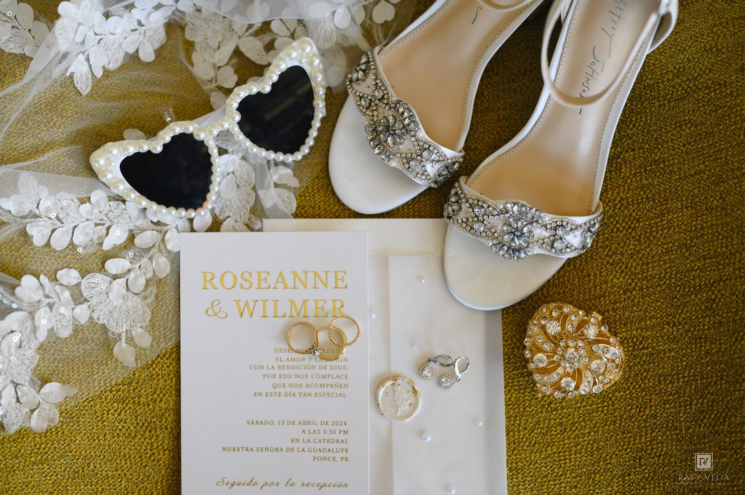 Wedding flat lay with rhinestone heels, heart sunglasses, floral lace, rings, and a Roseanne & Wilmer invitation on green.