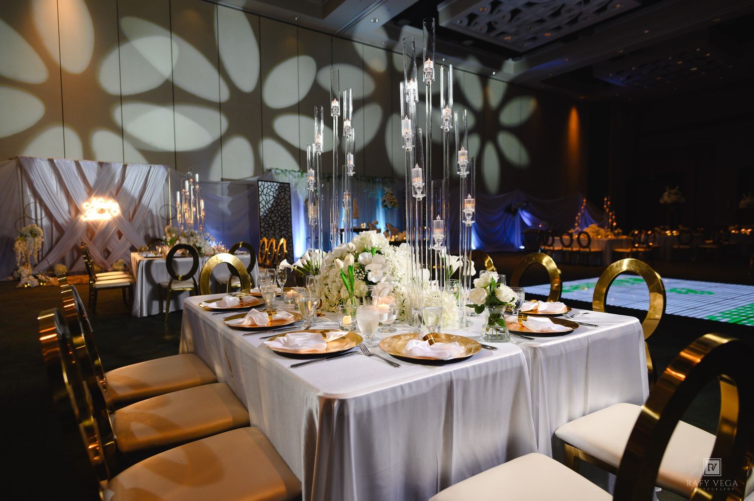 Elegant wedding reception table with crystal chandeliers, white floral centerpieces, gold chairs, and geometric wall projections.