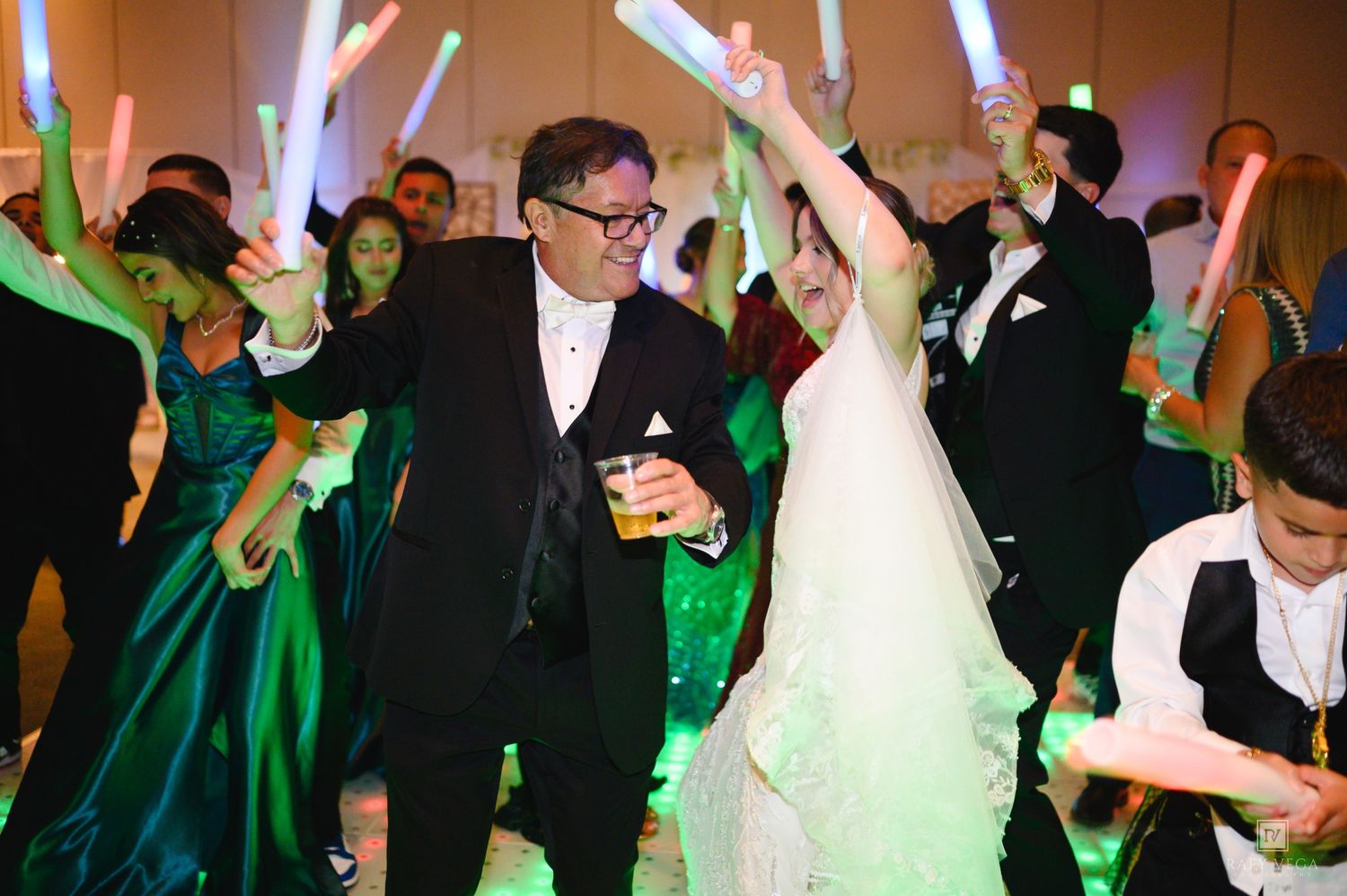 Bride and groom dancing joyfully at wedding reception with guests holding glowing light sticks in festive celebration.