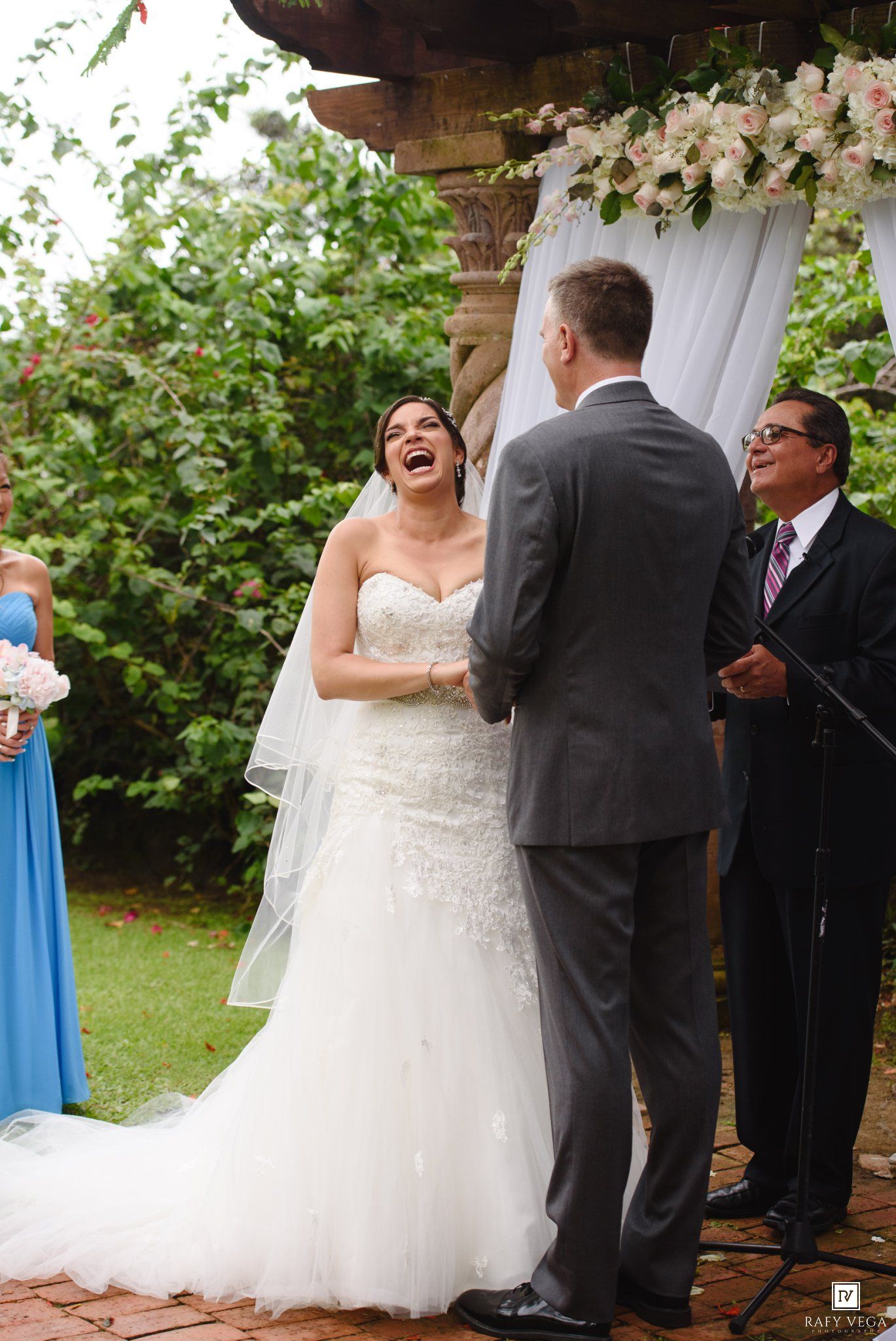Bride laughing joyfully during outdoor wedding ceremony in white lace gown, holding hands with groom in gray suit.