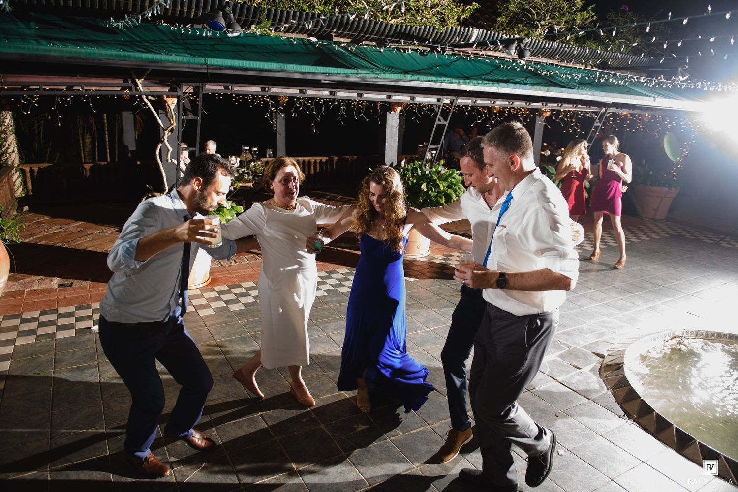 Wedding guests dance together on an outdoor patio at a festive evening reception with string lights.