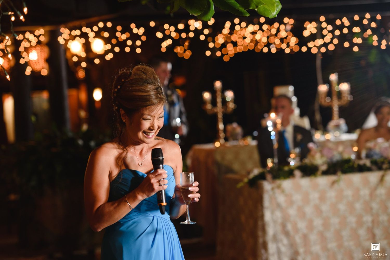 Woman in blue dress giving a toast speech at an elegant wedding reception with warm bokeh lights in the background.
