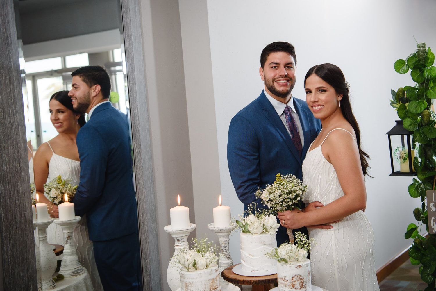 Bride and groom smiling beside wedding cake with candles and floral decor, reflected in a mirror.