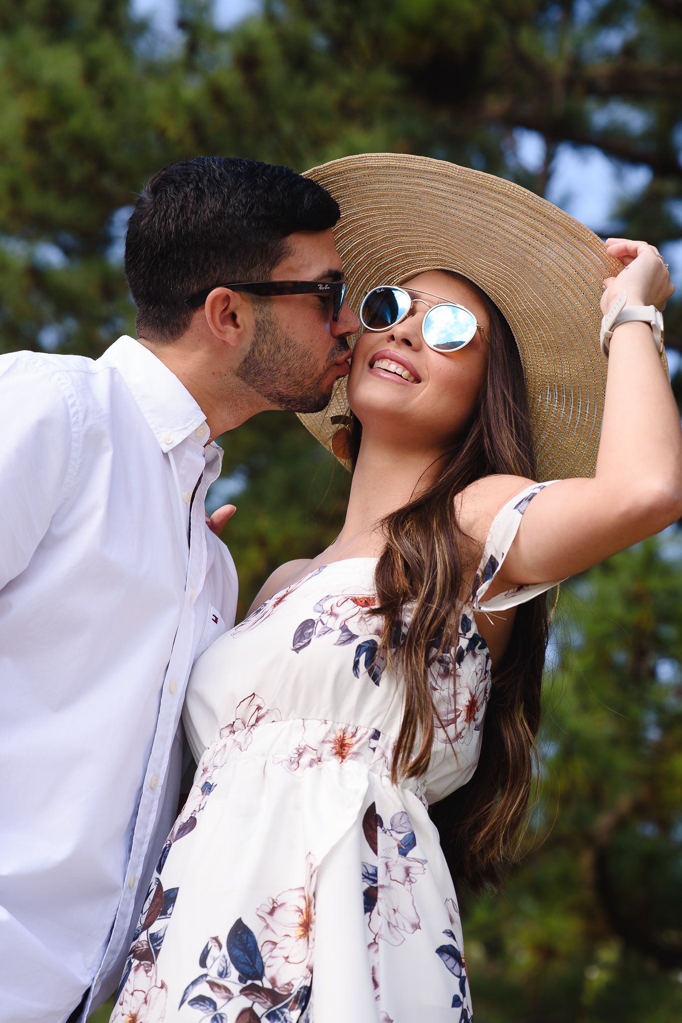 Romantic couple outdoors, man kissing woman's cheek, she wears floral dress, wide-brim hat, and mirrored sunglasses.
