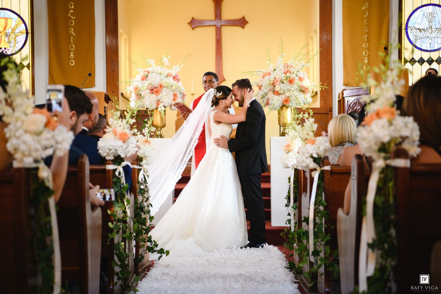 Bride and groom share first kiss at church altar adorned with white and pink floral arrangements during wedding ceremony.