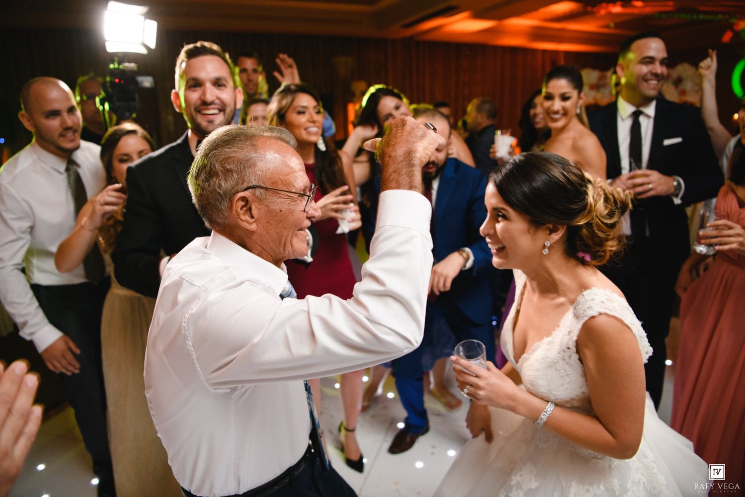 Bride and older man dancing joyfully at wedding reception surrounded by smiling guests in elegant venue.