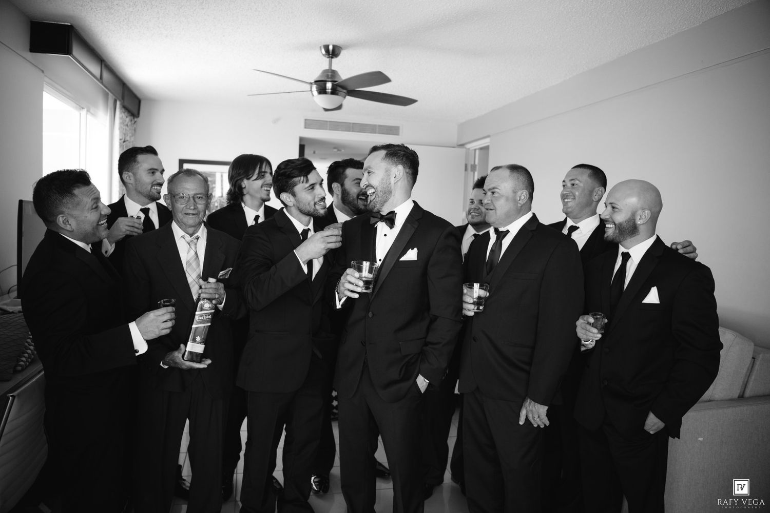 Groom and groomsmen in tuxedos sharing drinks and laughing together before wedding in black and white.